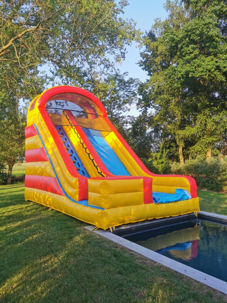 A large inflatable waterslide with yellow and red stripes set up beside a pool, surrounded by trees in a grassy area used ...