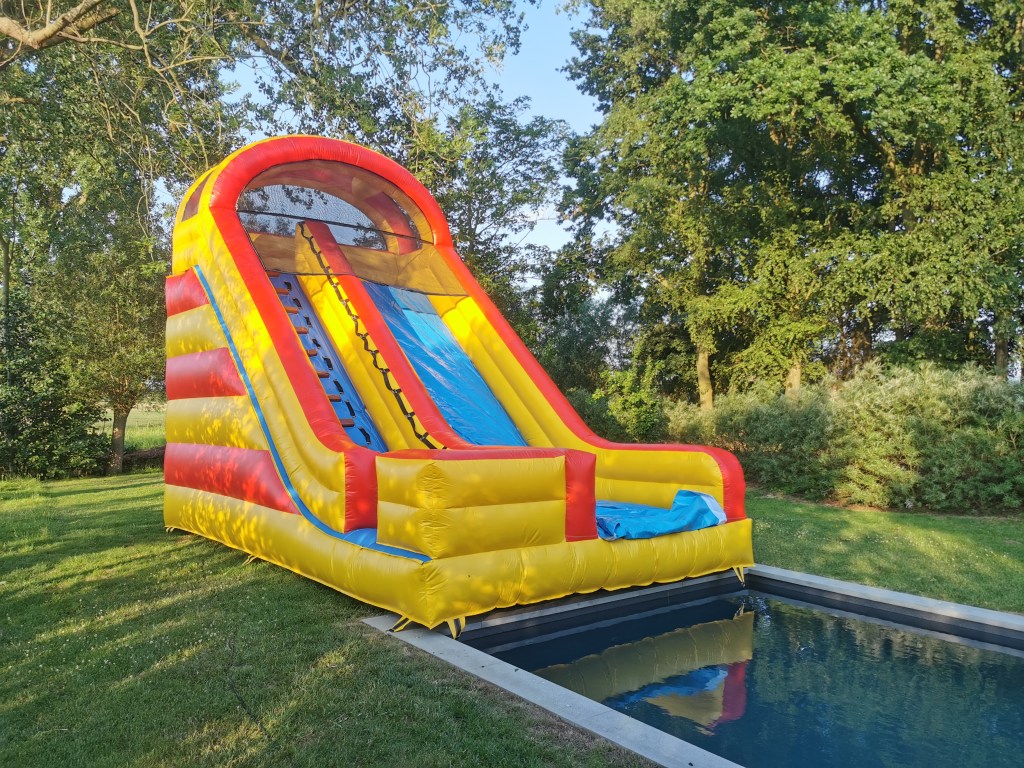 An inflatable slide, designed for kids, similar to a water slide, featured in a backyard with a rectangular, dark pool and...